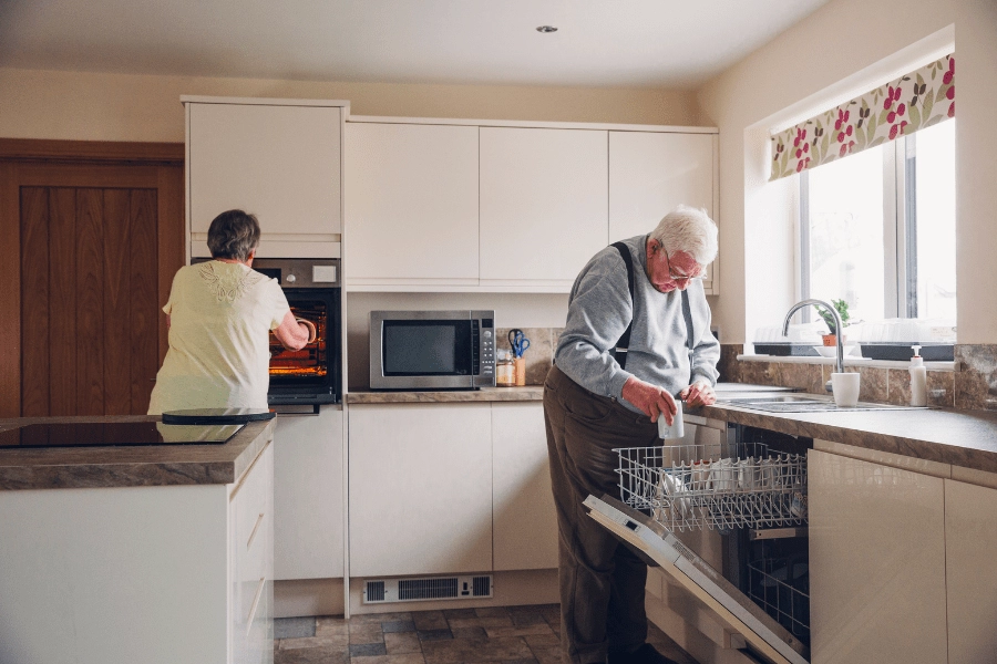 Senior woman taking food out of the oven while her elderly husband loads the dishwasher.