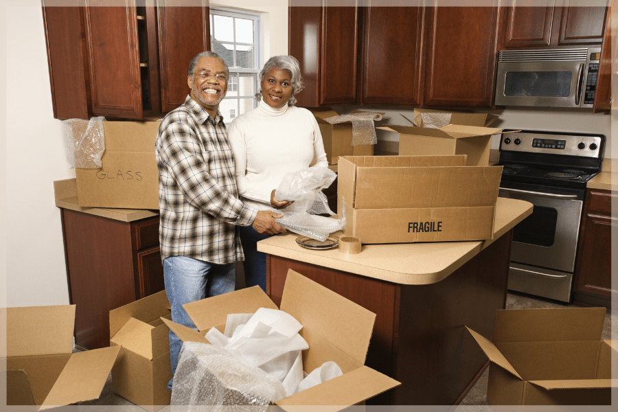 Downsizing for seniors - Smiling senior couple surrounded by bubble wrap and moving boxes in their kitchen - MeetCaregivers