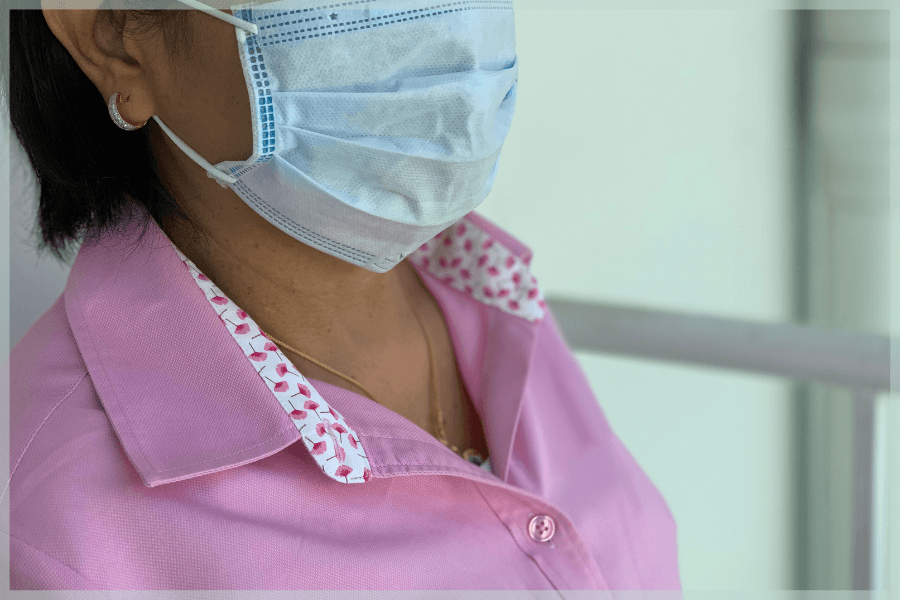 Social Isolation In Older Adults - Senior woman wearing a mask standing indoors beside a window - MeetCaregivers