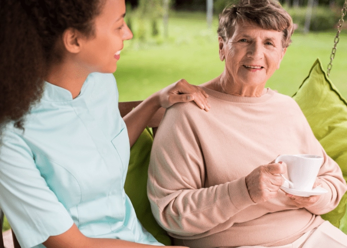 young caregiver and elderly woman holding tea sitting on an outdoor swing