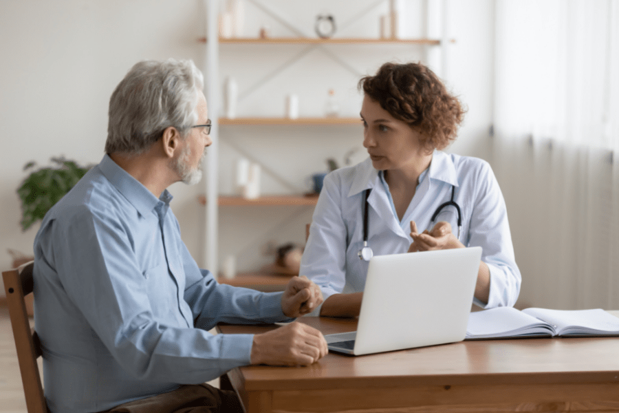 A doctor and middle aged man intensely discussing caring for elderly parents in front of a laptop and book