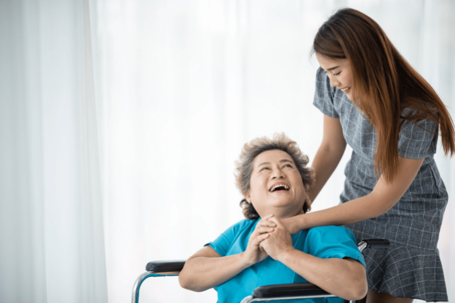 Certified Caregiver Standing Next To Her Laughing Grandma In A Wheelchair