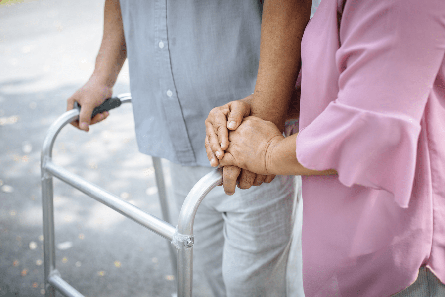 Balance Problems In Seniors - Woman clasping hands while walking outside with an elderly man with a walker
