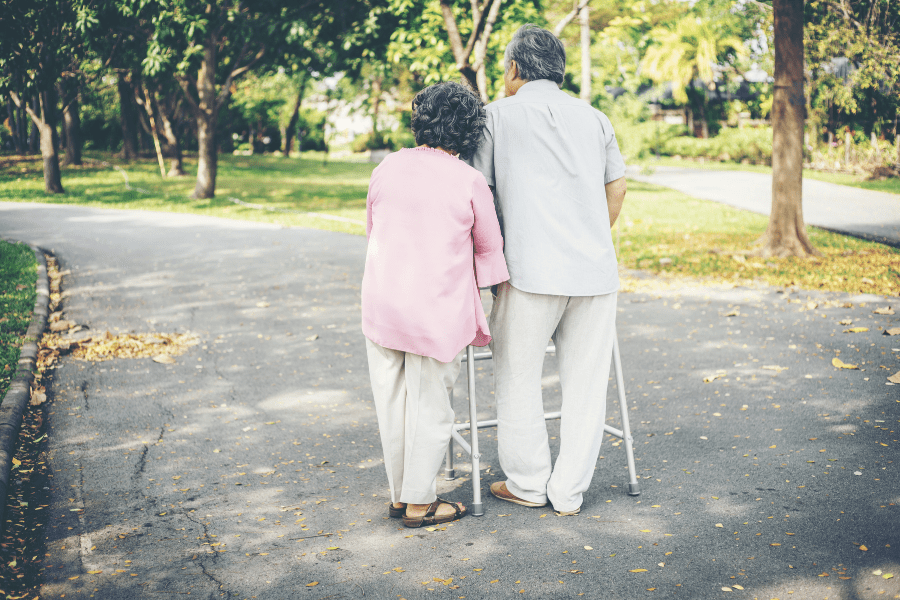 Elderly man using walker for support while walking outside with his wife.