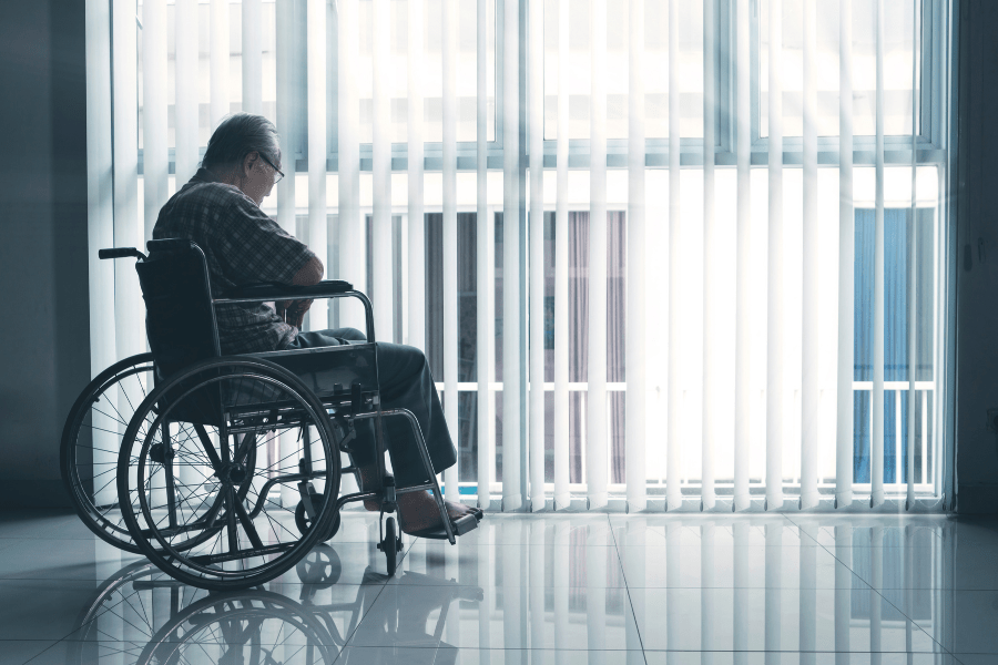Sad elderly man sitting in a wheelchair in front of a large window.